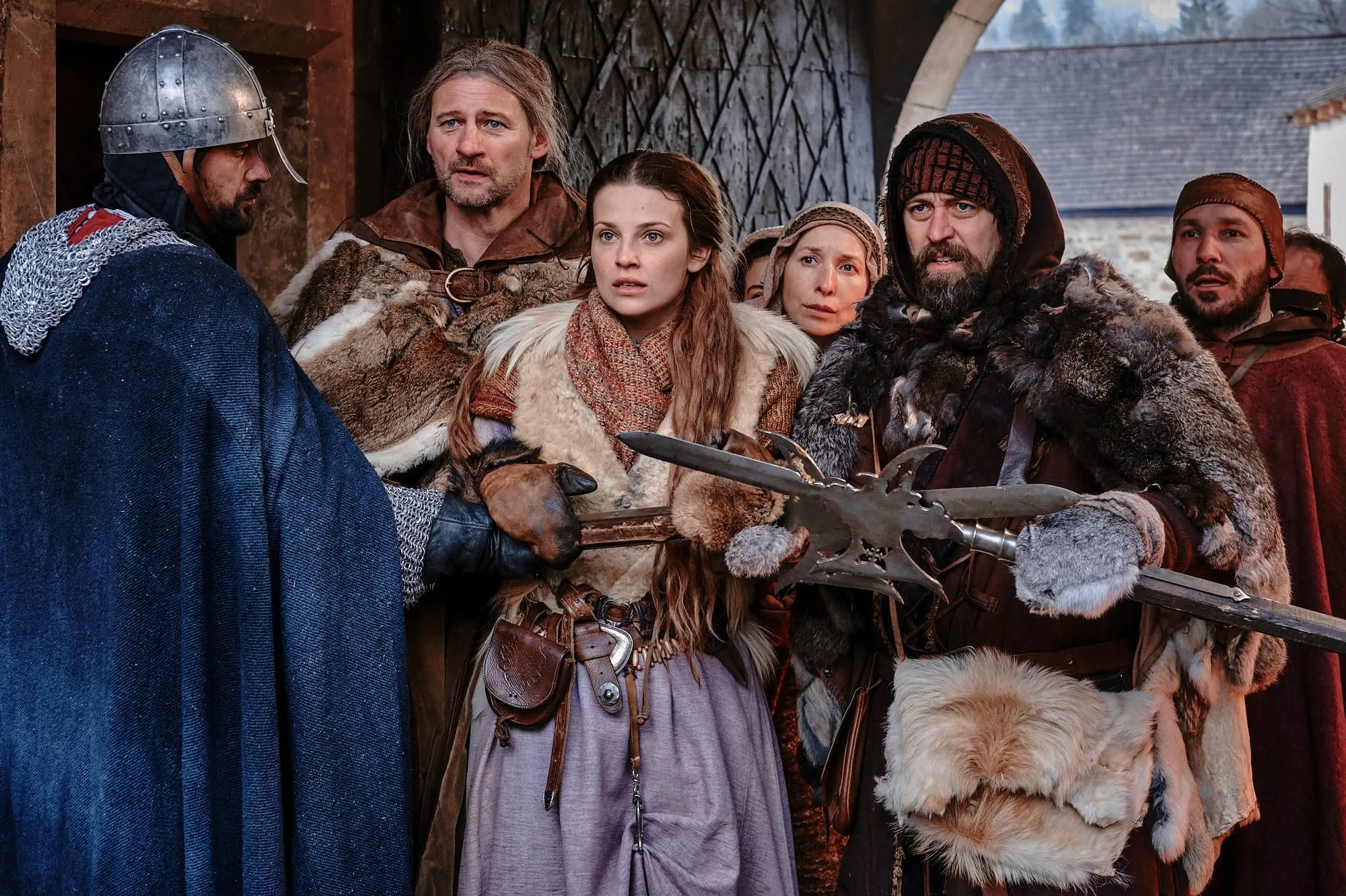 Villagers in furs face a helmeted soldier in chain mail at a gate; a bearded villager holds a halberd out in front of him