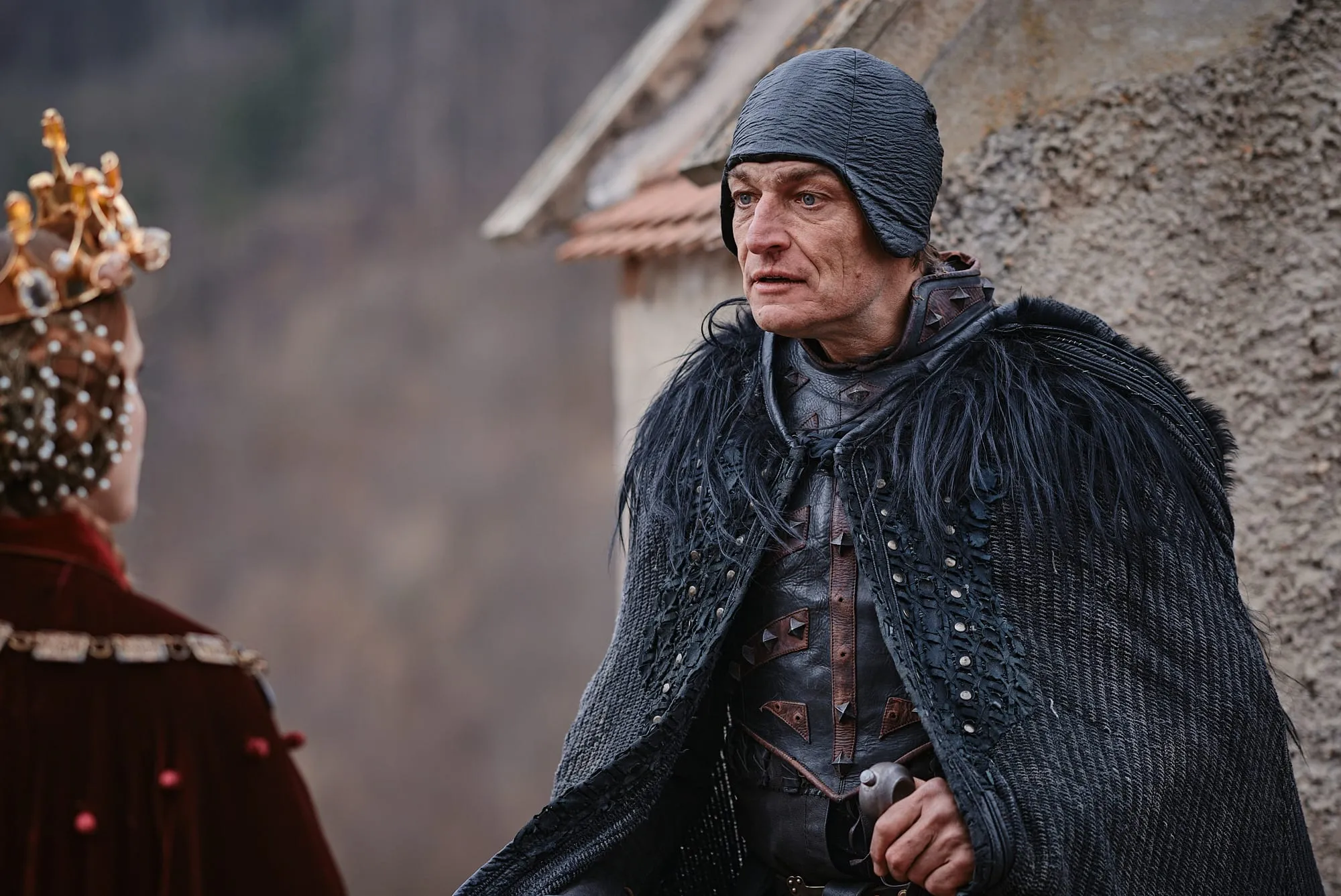 A man in a black feathered cloak speaks to a woman in a golden crown who has her back to the camera