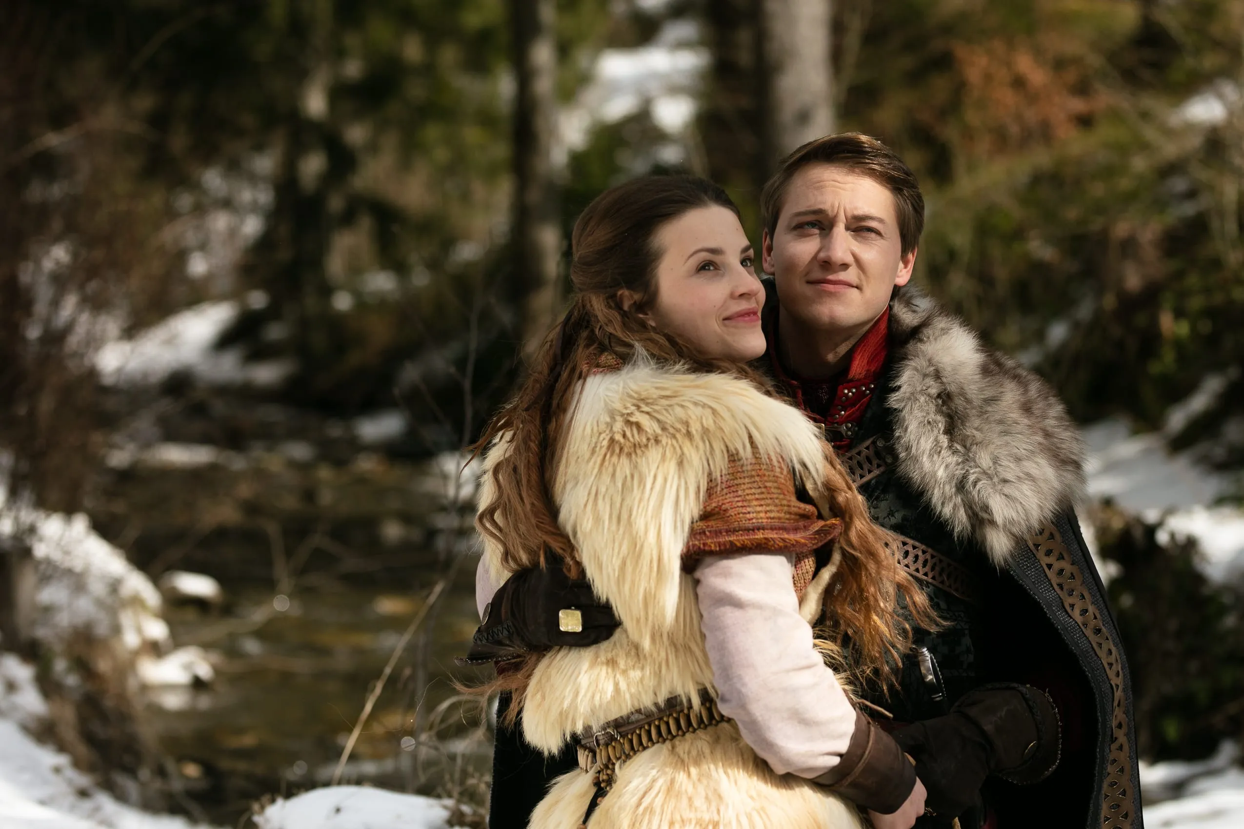 A young couple embrace in a snowy forest beside a stream, both smiling and looking upwards