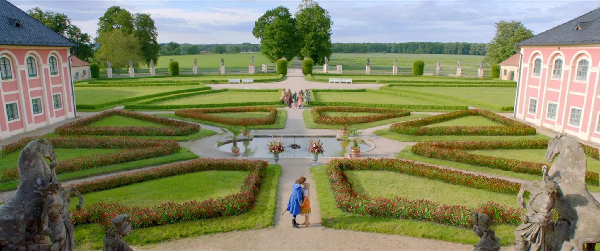 A formal palace garden with clipped hedges; in the foreground a man in a blue coat walks with a girl in an orange dress