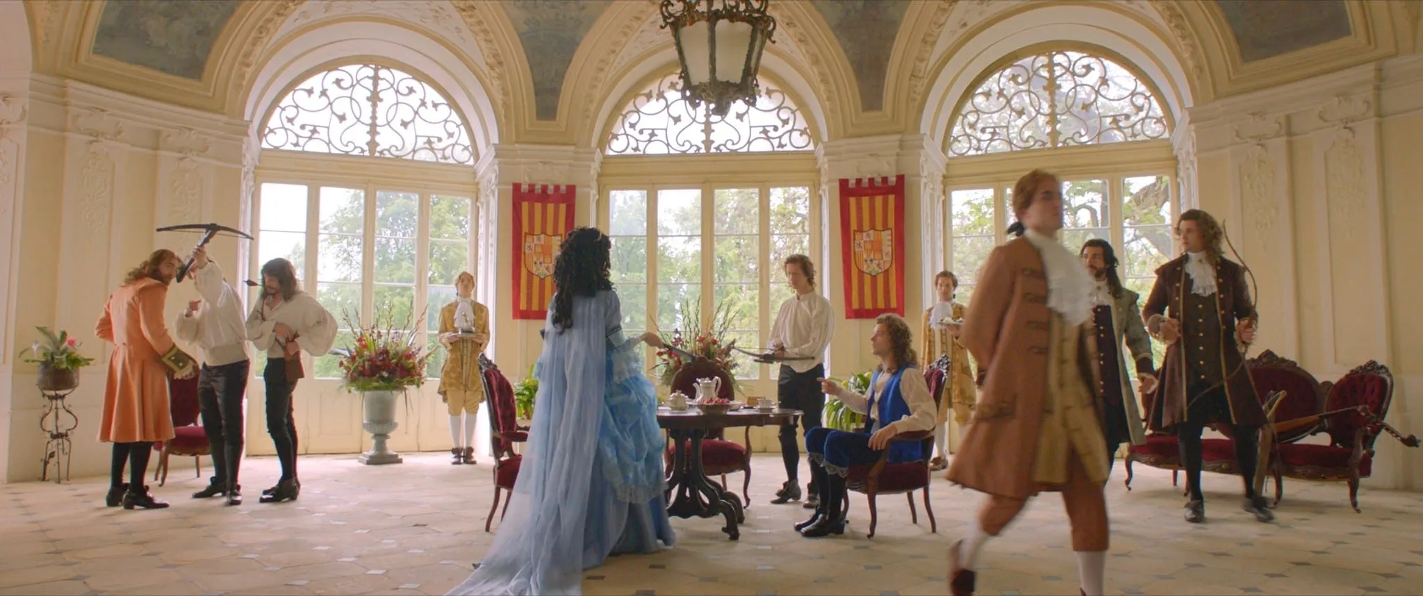 In a bright palace hall a woman in a pale blue gown stands with her back to the camera, surrounded by courtiers in Baroque coats