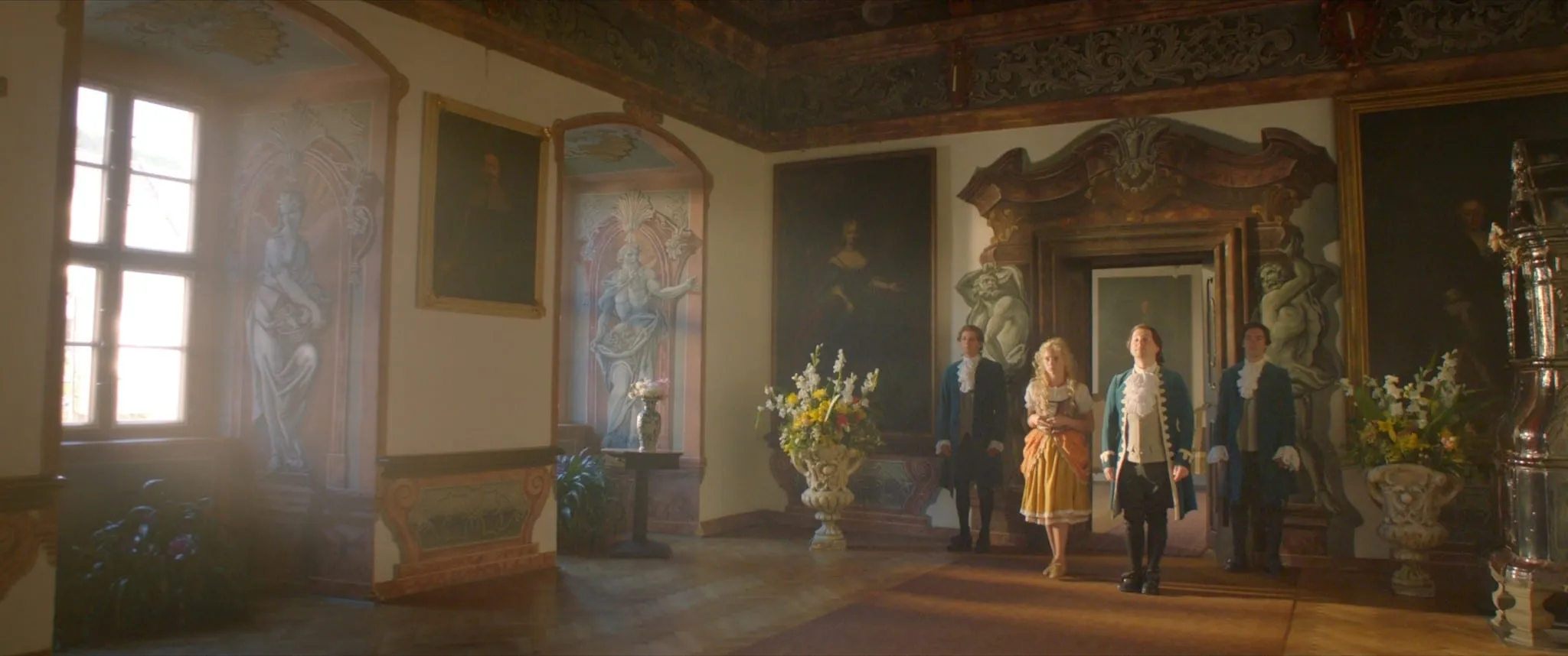 In a frescoed hall a fair-haired girl in an orange dress stands among three men in blue period coats