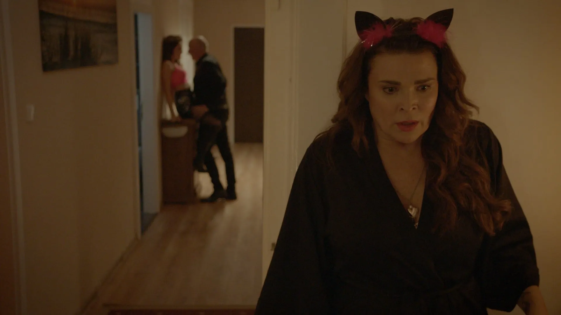 Close-up of a woman in a black dressing gown and a cat-ear headband looking sideways down a corridor in dismay