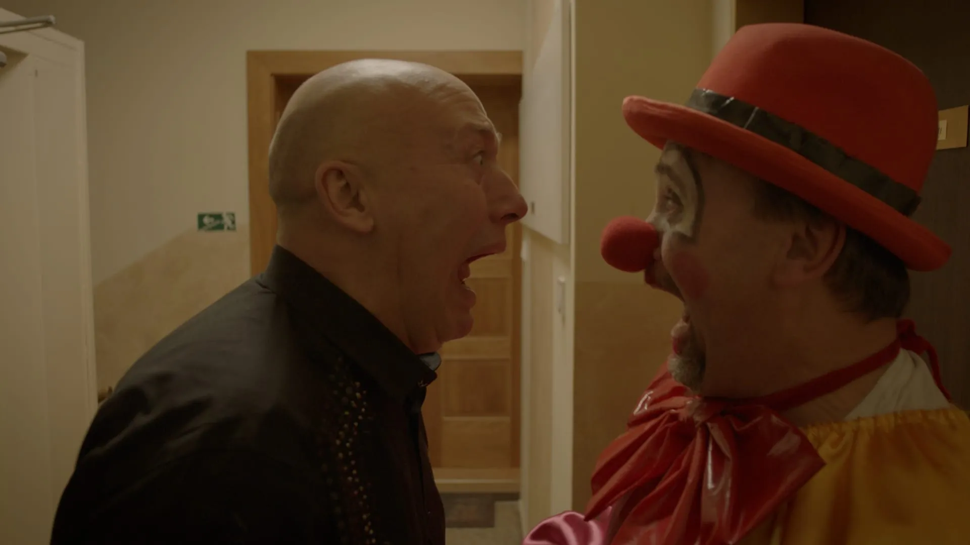 A bald man in a black shirt shouts right in the face of a clown in a red bowler hat with a red nose