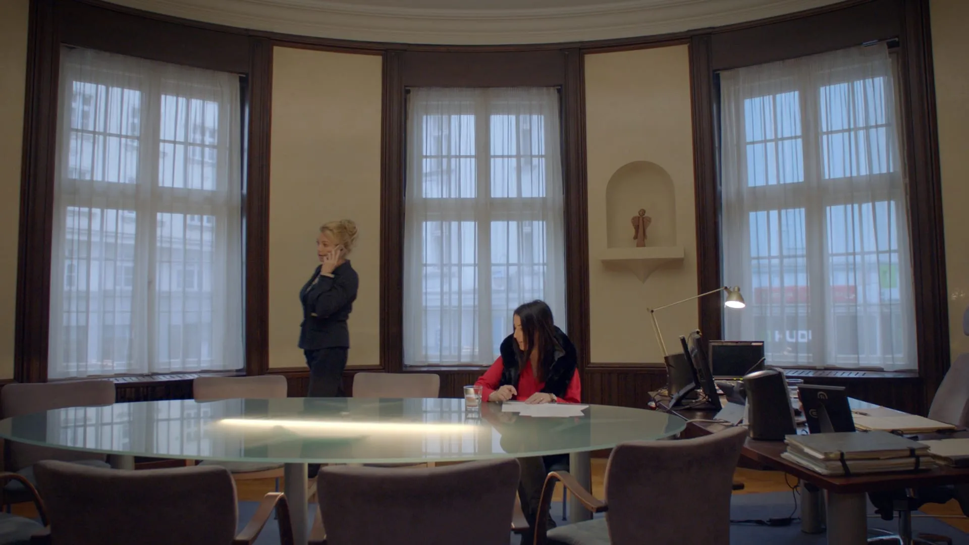 In an office with tall windows a blonde woman in a black suit takes a call while a dark-haired woman in red signs papers at a desk