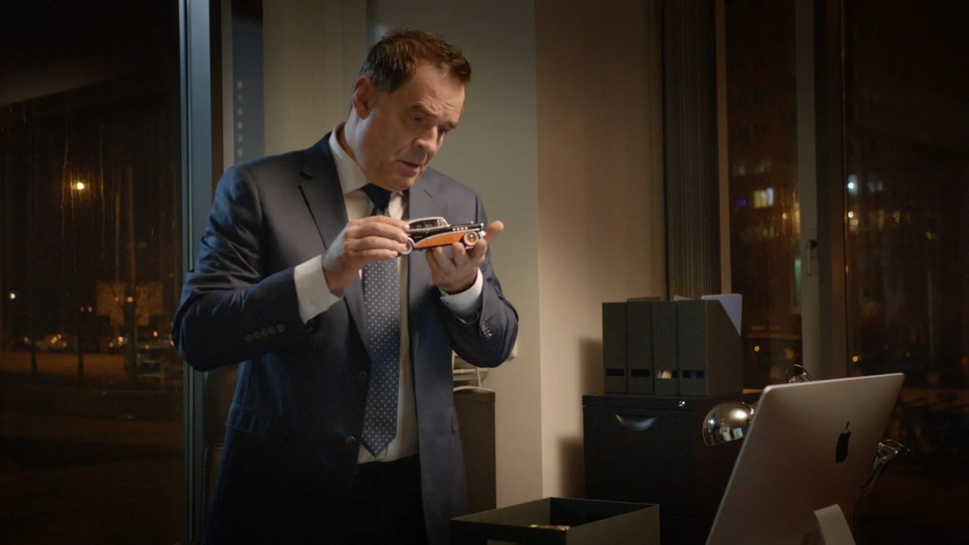 A man in a blue suit stands by an office window in the evening, examining a small model car in his hands