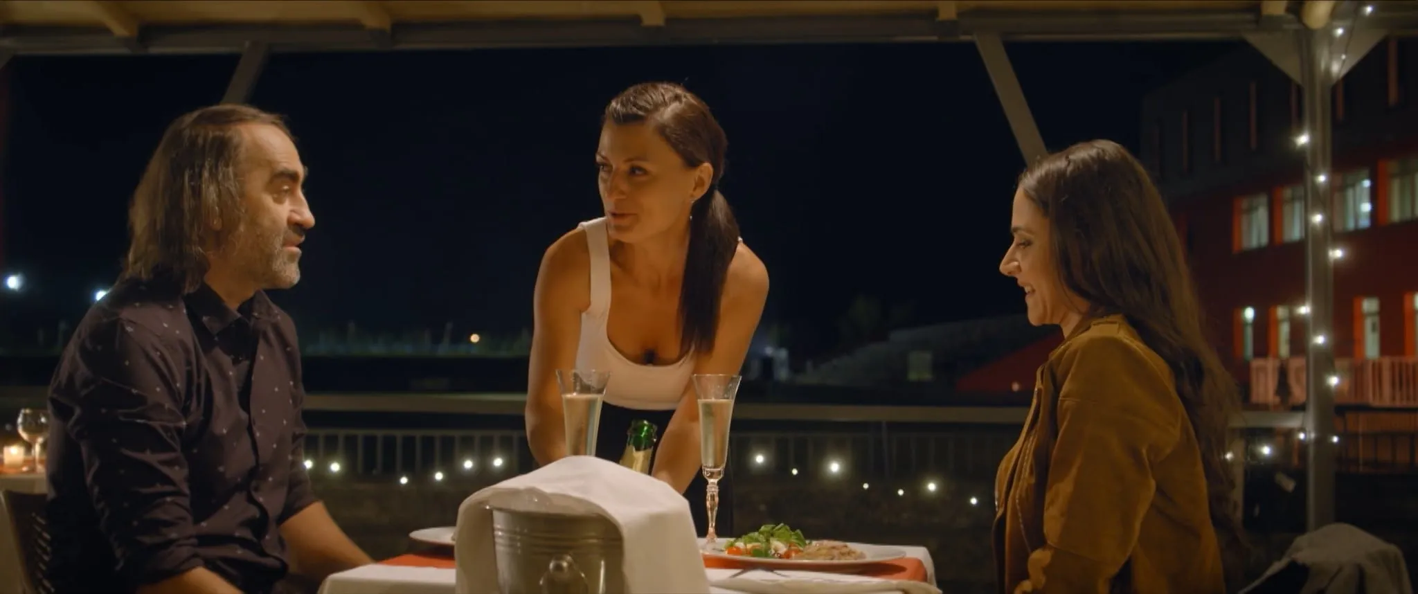 At a table on a terrace in the evening a long-haired man looks at a waitress in a white top setting down champagne glasses