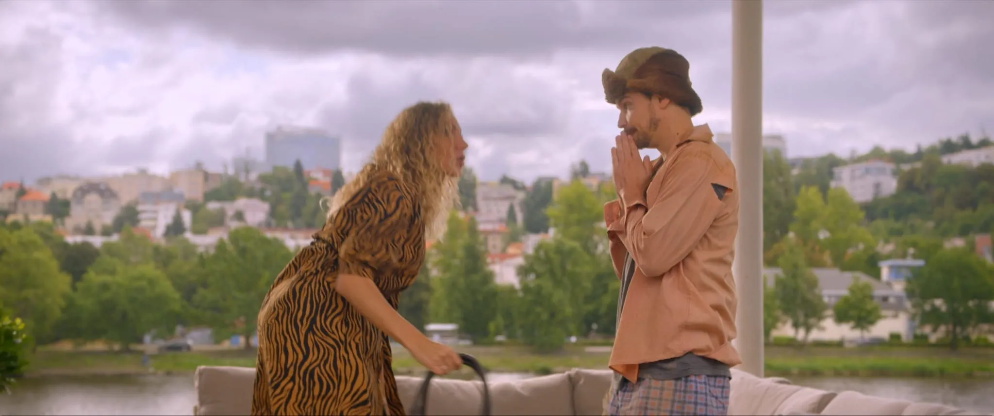 A blonde woman in a tiger-print dress leans over a sofa towards a man in a fur hat who presses his palms together, pleading
