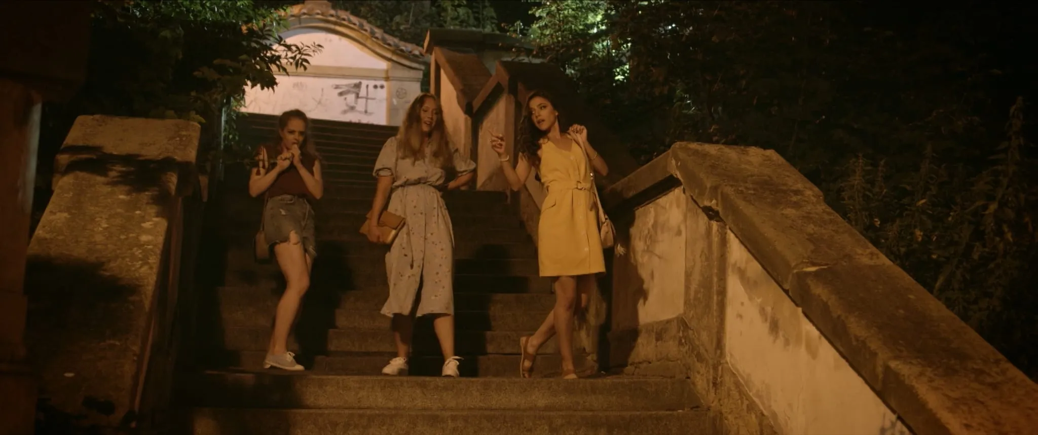 Three young women in summer dresses come down stone steps at night, the one in a yellow dress gesturing with a cigarette