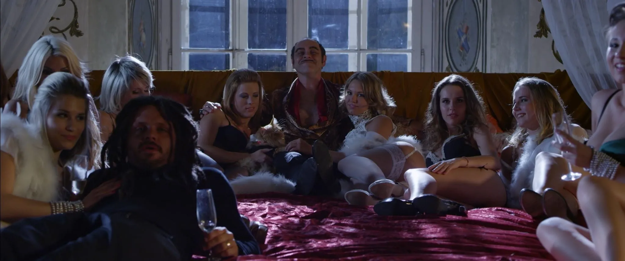 A man in a silk robe sits on a wide bed surrounded by women in lingerie, tall windows behind him