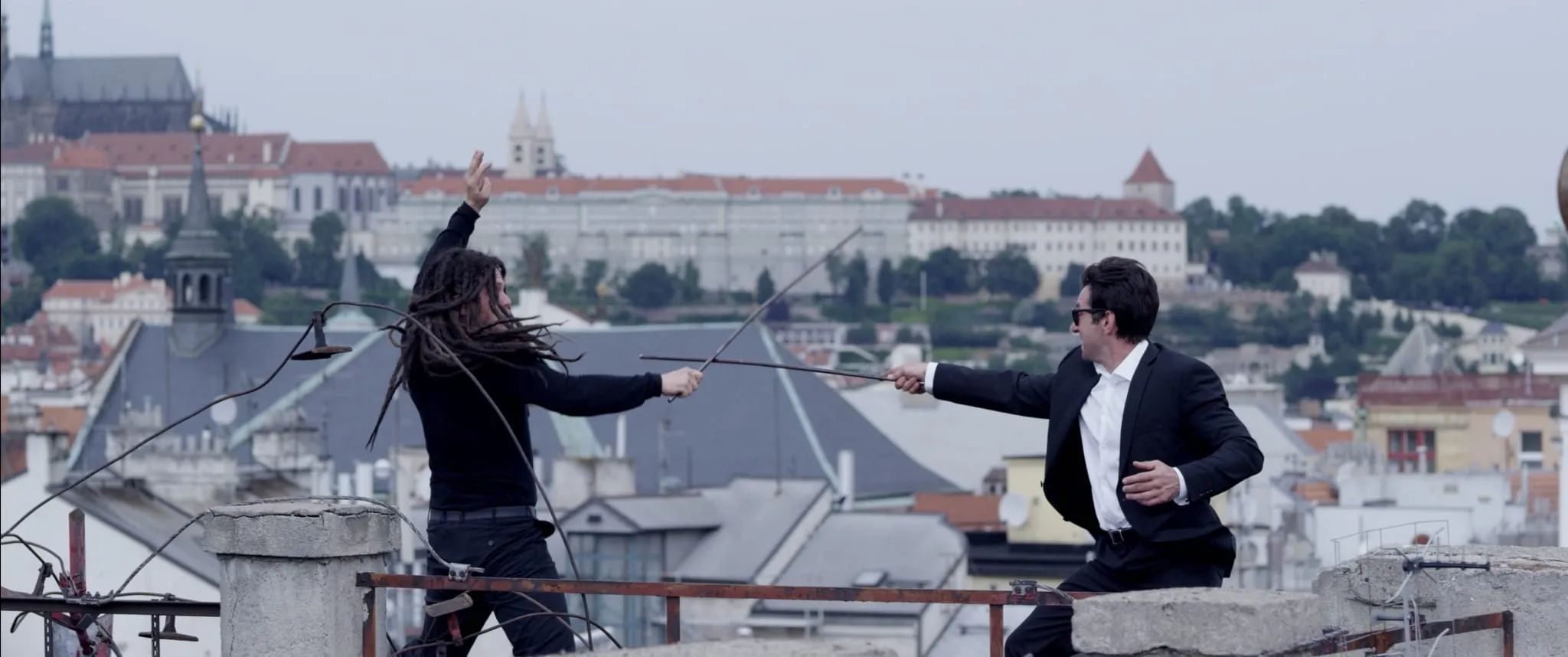 Two men fence with thin rods on a rooftop, Prague Castle outlined in the background